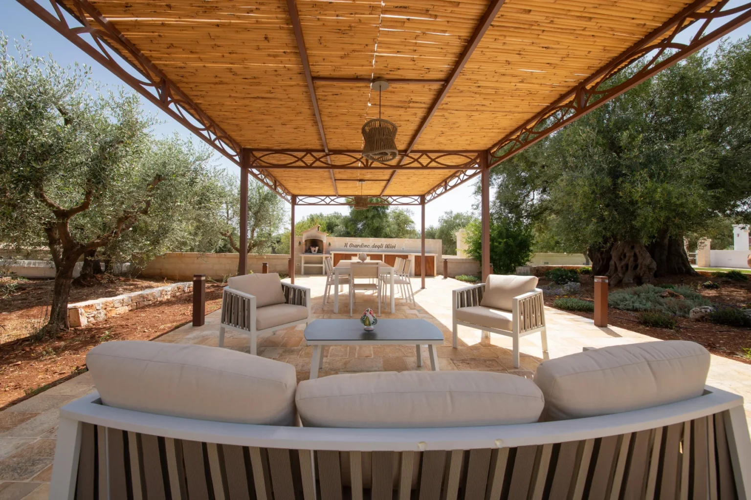 Outdoor seating area under a wooden pergola, featuring comfortable chairs and a table, surrounded by olive trees and a stone path, ideal for relaxation and gatherings. | Only on SopranoVillas