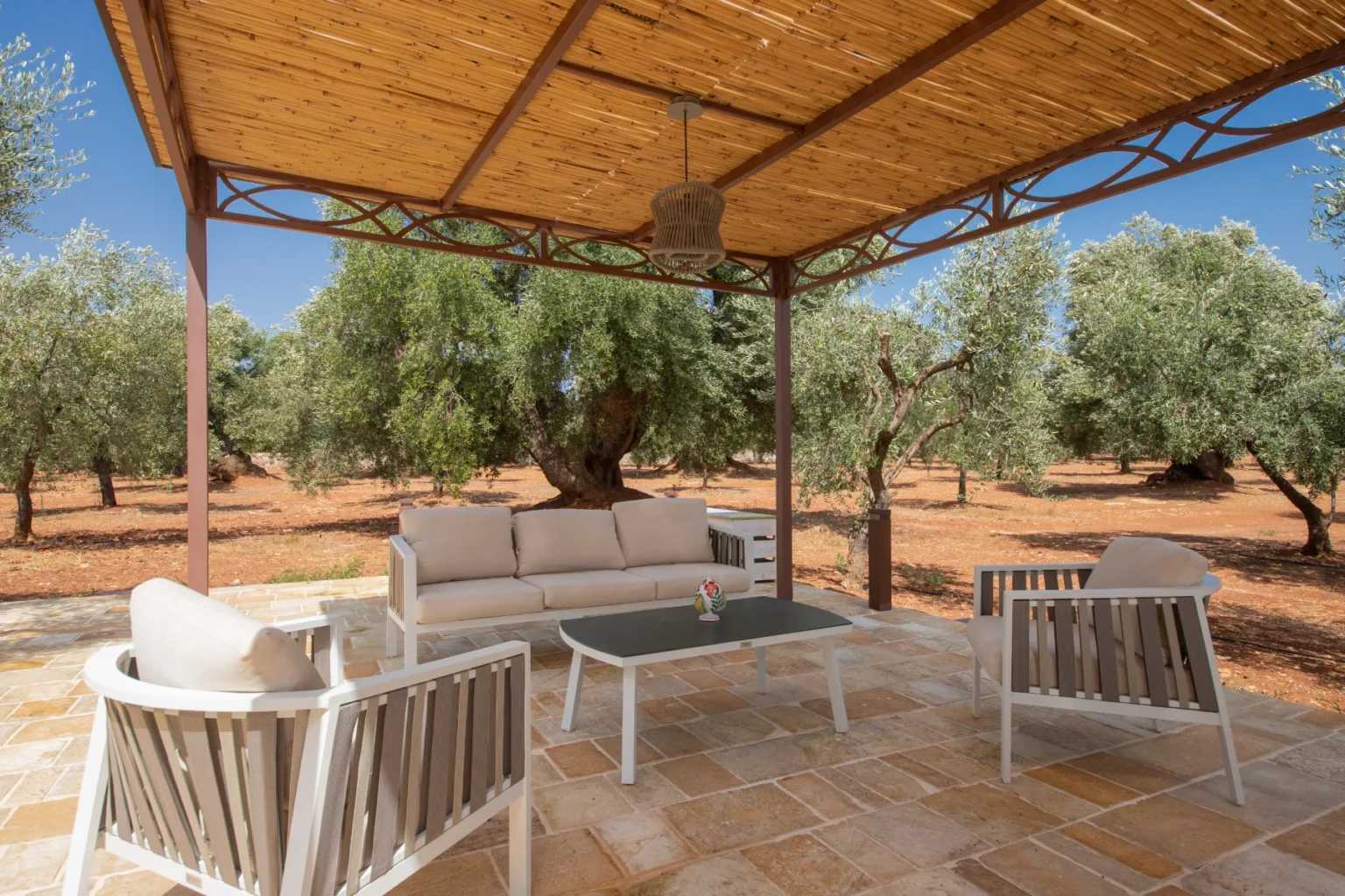 Outdoor seating area under a wooden pergola, featuring a light-colored sofa and chairs, surrounded by olive trees in a serene landscape. Ideal for relaxation and enjoying nature. | Only on SopranoVillas