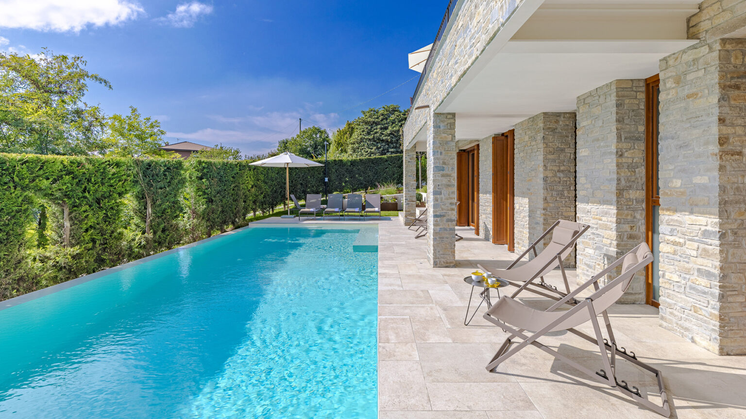 Luxury villa with a sparkling blue swimming pool and modern patio seating, surrounded by lush greenery and clear blue skies. Ideal for relaxation and outdoor leisure. | Only on SopranoVillas