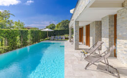Luxury villa with a sparkling blue swimming pool and modern patio seating, surrounded by lush greenery and clear blue skies. Ideal for relaxation and outdoor leisure.