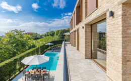 Modern villa exterior with a swimming pool, patio furniture, and lush greenery under a clear blue sky. Ideal for luxury living and outdoor relaxation.