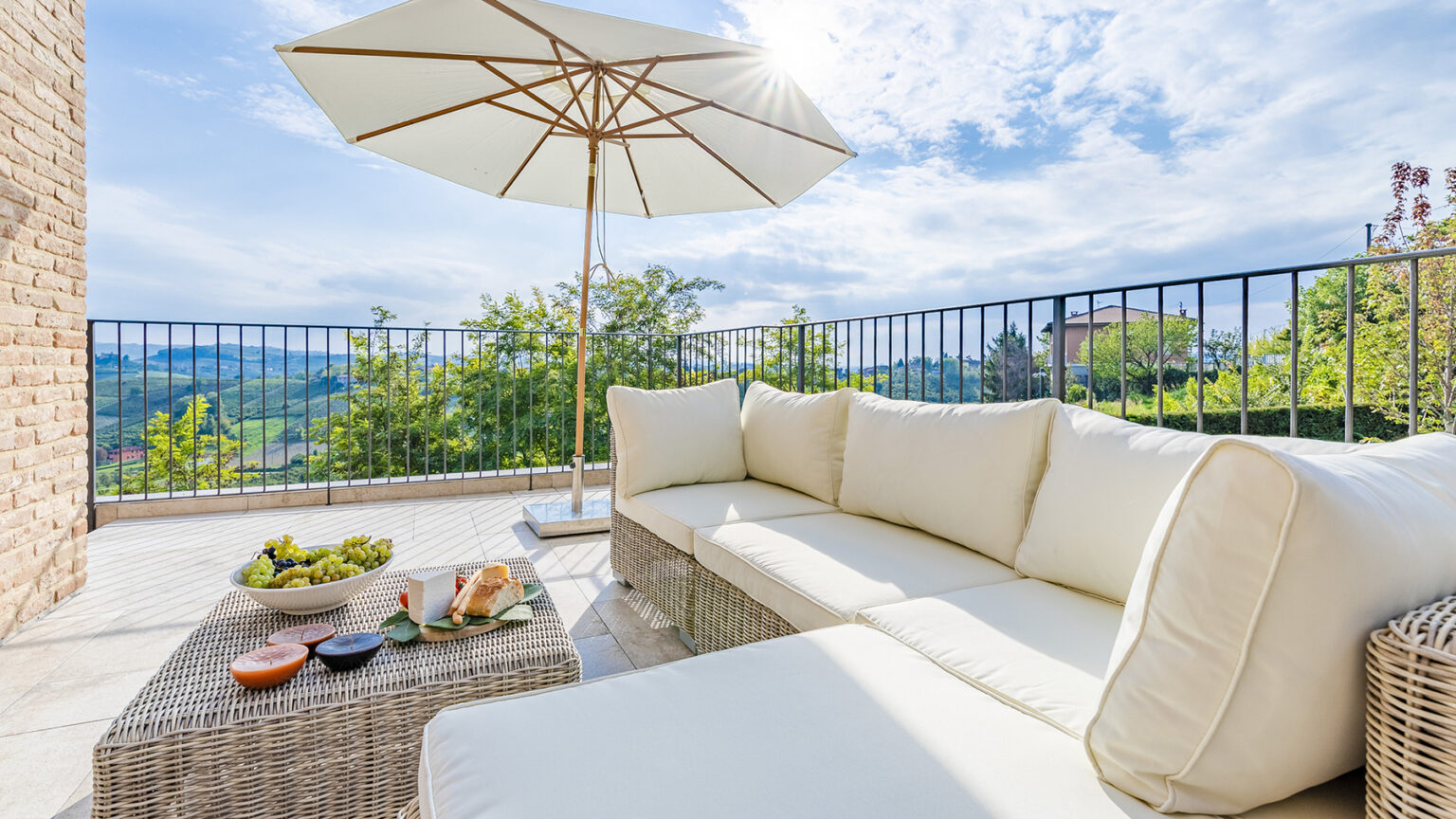Cozy outdoor seating area featuring a white sectional sofa, a wicker coffee table with snacks, and a large umbrella for shade, set against a scenic landscape with greenery and a clear blue sky. | Only on SopranoVillas
