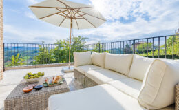 Cozy outdoor seating area featuring a white sectional sofa, a wicker coffee table with snacks, and a large umbrella for shade, set against a scenic landscape with greenery and a clear blue sky.
