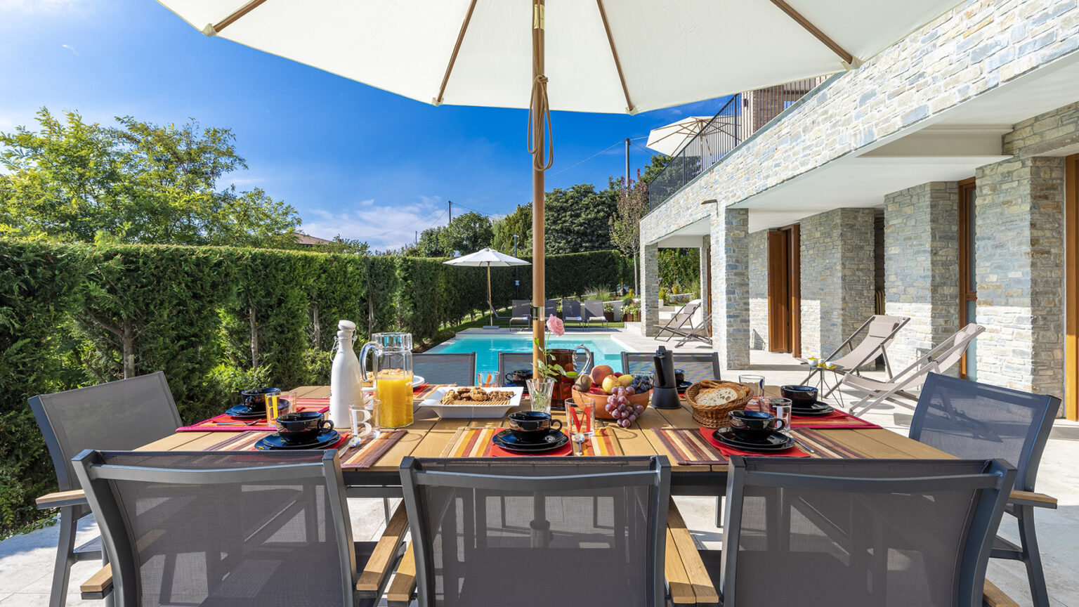 Outdoor dining area with a wooden table set for breakfast, featuring plates, glasses, and a variety of drinks, under a large umbrella. In the background, a swimming pool and lush greenery create a relaxing atmosphere. | Only on SopranoVillas