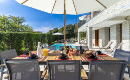 Outdoor dining area with a wooden table set for breakfast, featuring plates, glasses, and a variety of drinks, under a large umbrella. In the background, a swimming pool and lush greenery create a relaxing atmosphere.