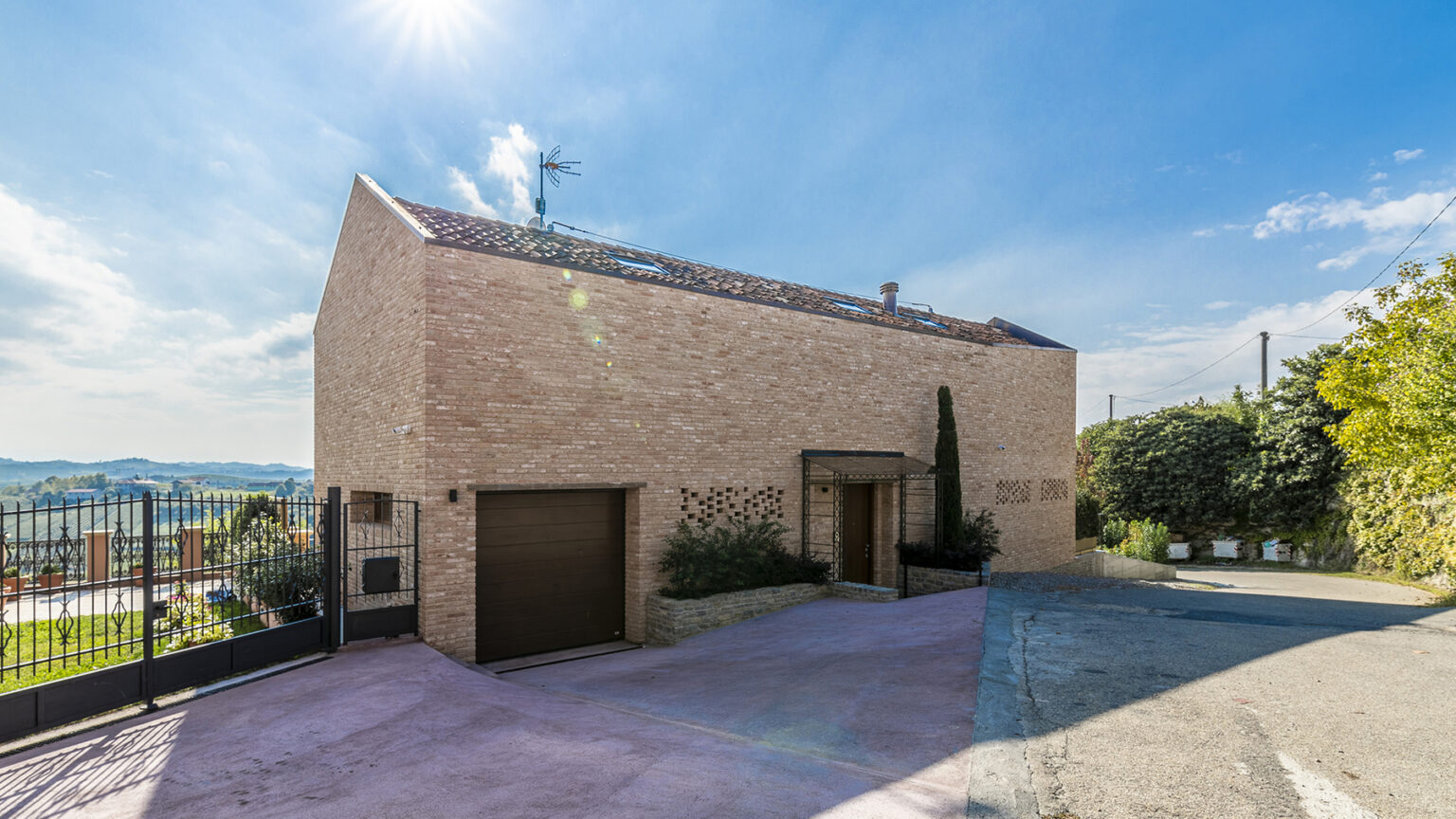 Modern residential building with a minimalist design, featuring a brick facade, large windows, and a garage, set against a clear blue sky and green landscape. | Only on SopranoVillas