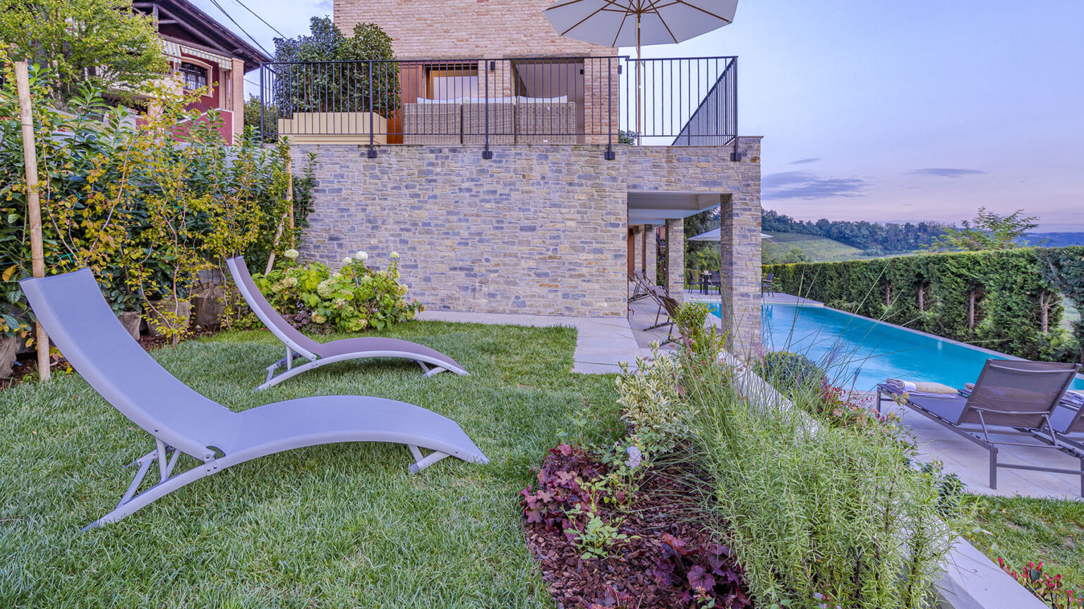 Luxury outdoor area featuring two lounge chairs on a manicured lawn, a modern stone wall, and a swimming pool. The scene includes lush greenery and a patio with an umbrella, ideal for relaxation and leisure. | Only on SopranoVillas