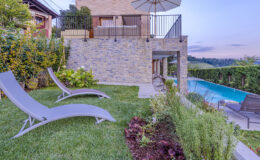 Luxury outdoor area featuring two lounge chairs on a manicured lawn, a modern stone wall, and a swimming pool. The scene includes lush greenery and a patio with an umbrella, ideal for relaxation and leisure.