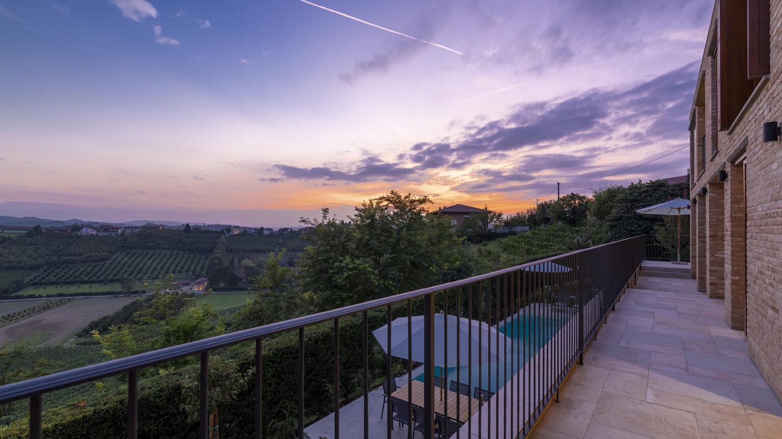 Scenic view from a balcony overlooking lush vineyards at sunset, with a tranquil sky and modern architecture in the foreground. | Only on SopranoVillas