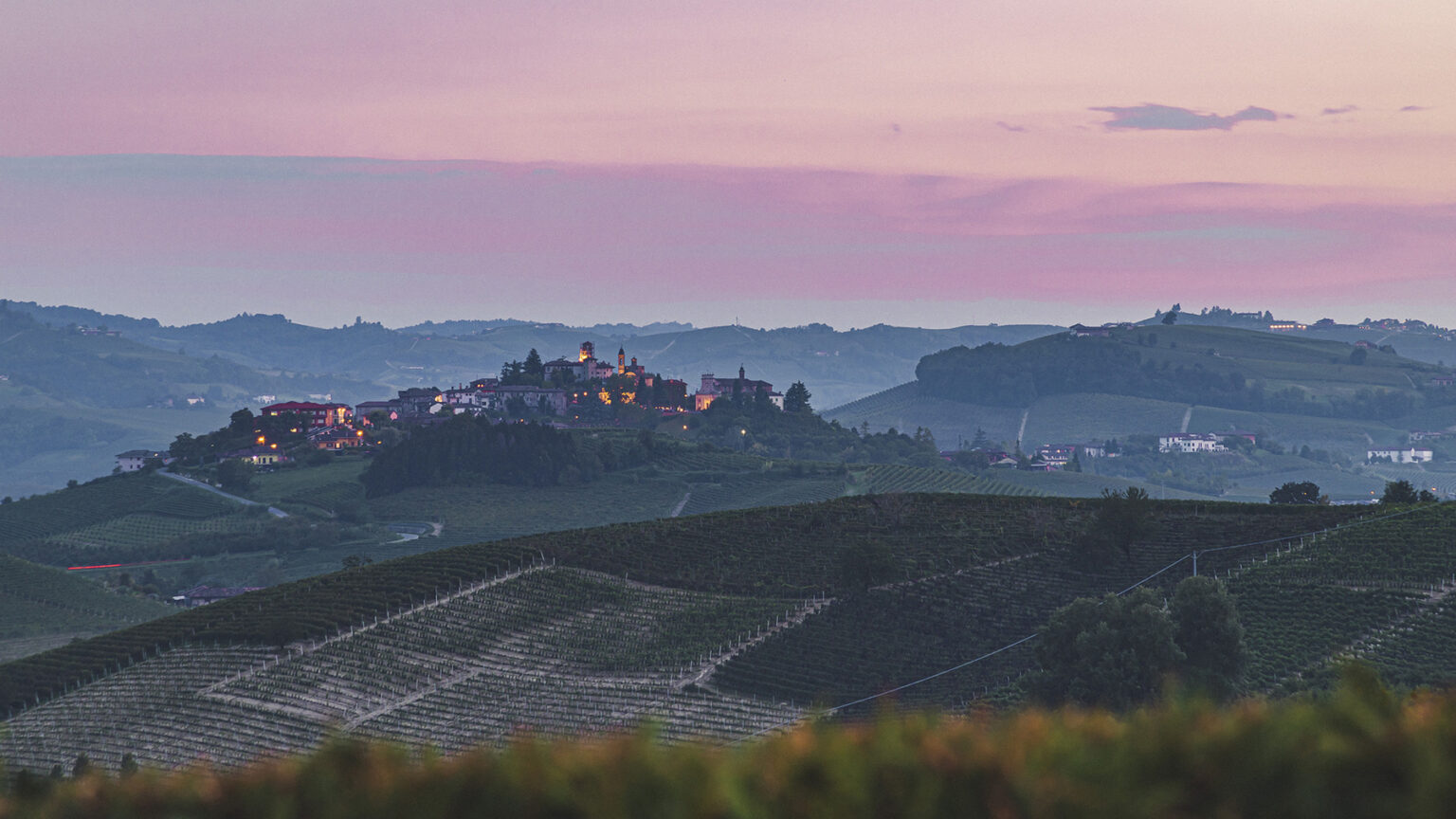 Scenic view of a hillside village illuminated at dusk, surrounded by rolling vineyards and a colorful sky in the background, showcasing the beauty of the countryside. | Only on SopranoVillas