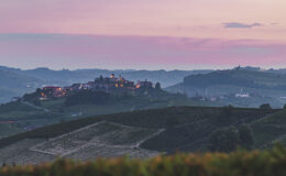 Scenic view of a hillside village illuminated at dusk, surrounded by rolling vineyards and a colorful sky in the background, showcasing the beauty of the countryside.