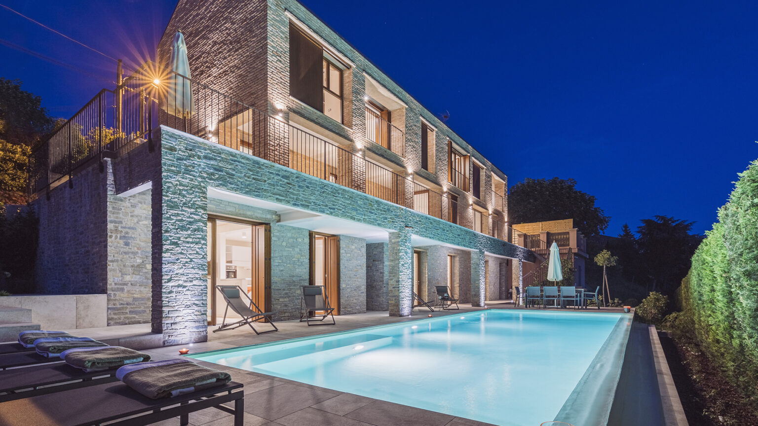 Modern villa with a swimming pool at night, featuring elegant architecture and ambient lighting, surrounded by lush greenery. | Only on SopranoVillas