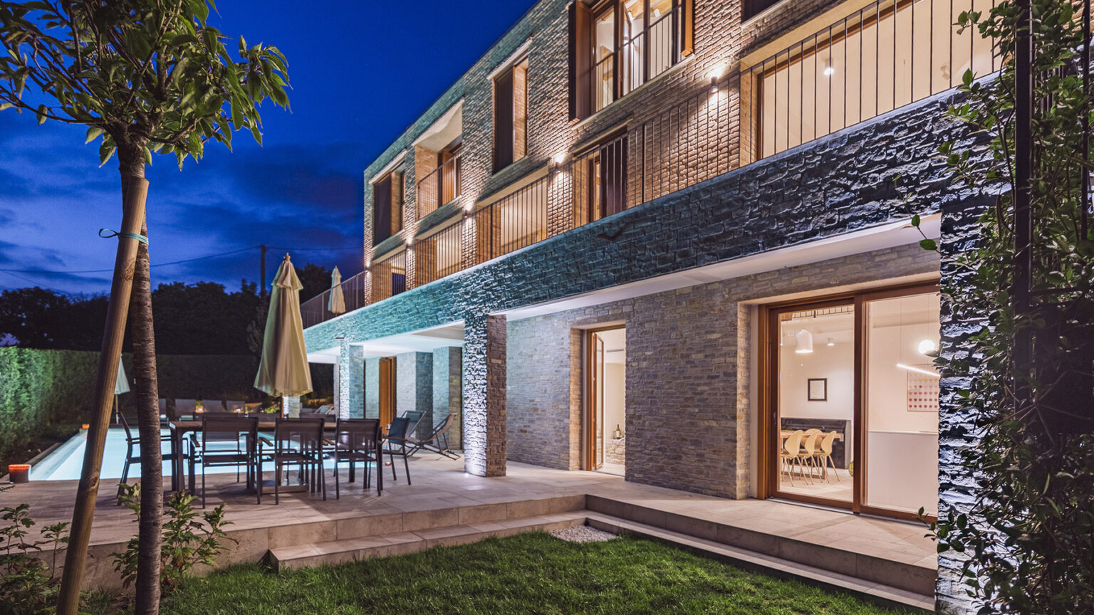 Modern residential home exterior with a stone facade, outdoor seating area, and illuminated pool at dusk. Lush green landscaping and patio create a serene atmosphere. | Only on SopranoVillas