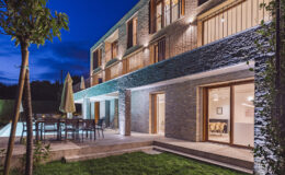 Modern residential home exterior with a stone facade, outdoor seating area, and illuminated pool at dusk. Lush green landscaping and patio create a serene atmosphere.