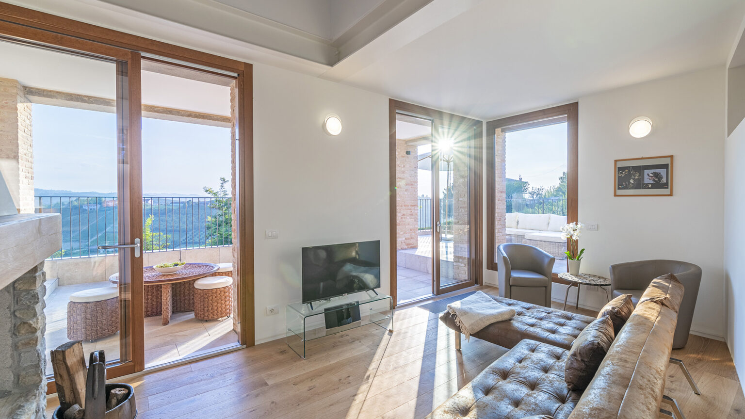 Modern living room with large windows, wooden flooring, and stylish furniture, featuring a cozy seating area and a view of an outdoor terrace. Natural light enhances the inviting atmosphere. | Only on SopranoVillas