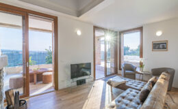 Modern living room with large windows, wooden flooring, and stylish furniture, featuring a cozy seating area and a view of an outdoor terrace. Natural light enhances the inviting atmosphere.
