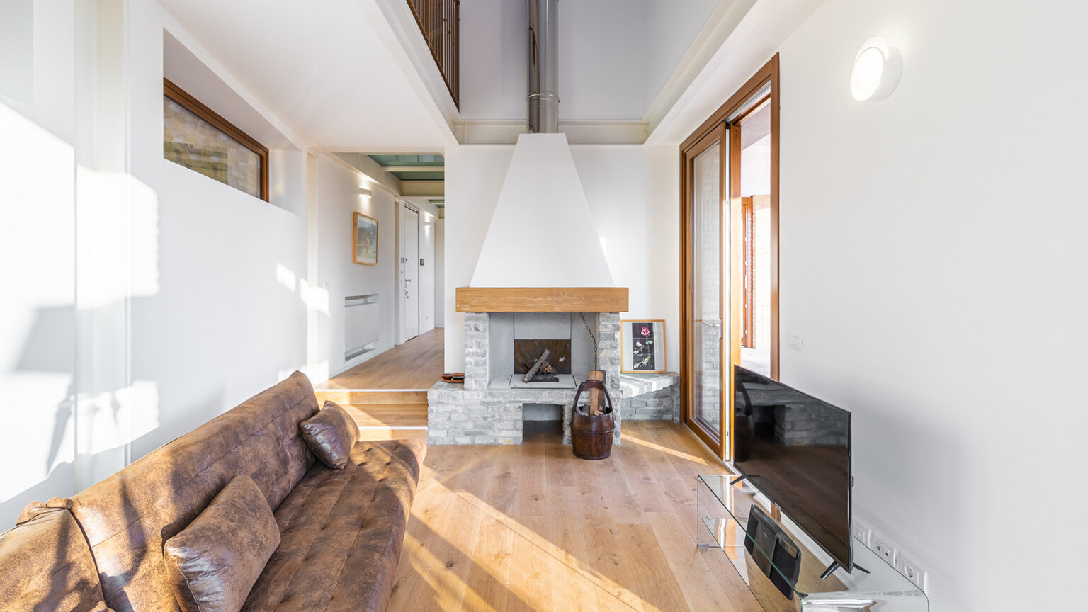 Modern living room with a cozy brown sofa, stone fireplace, and large windows allowing natural light to fill the space. Minimalist decor with wooden flooring and a flat-screen TV enhances the inviting atmosphere. | Only on SopranoVillas