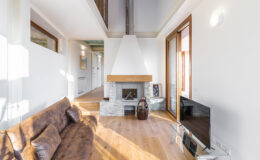 Modern living room with a cozy brown sofa, stone fireplace, and large windows allowing natural light to fill the space. Minimalist decor with wooden flooring and a flat-screen TV enhances the inviting atmosphere.
