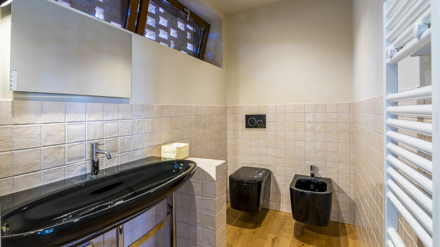 Modern bathroom interior featuring a sleek black sink, two black toilets, and tiled walls with a warm wooden floor. Natural light filters through unique window designs, enhancing the contemporary aesthetic. Ideal for showcasing stylish bathroom design ideas. | Only on SopranoVillas