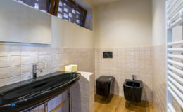 Modern bathroom interior featuring a sleek black sink, two black toilets, and tiled walls with a warm wooden floor. Natural light filters through unique window designs, enhancing the contemporary aesthetic. Ideal for showcasing stylish bathroom design ideas.