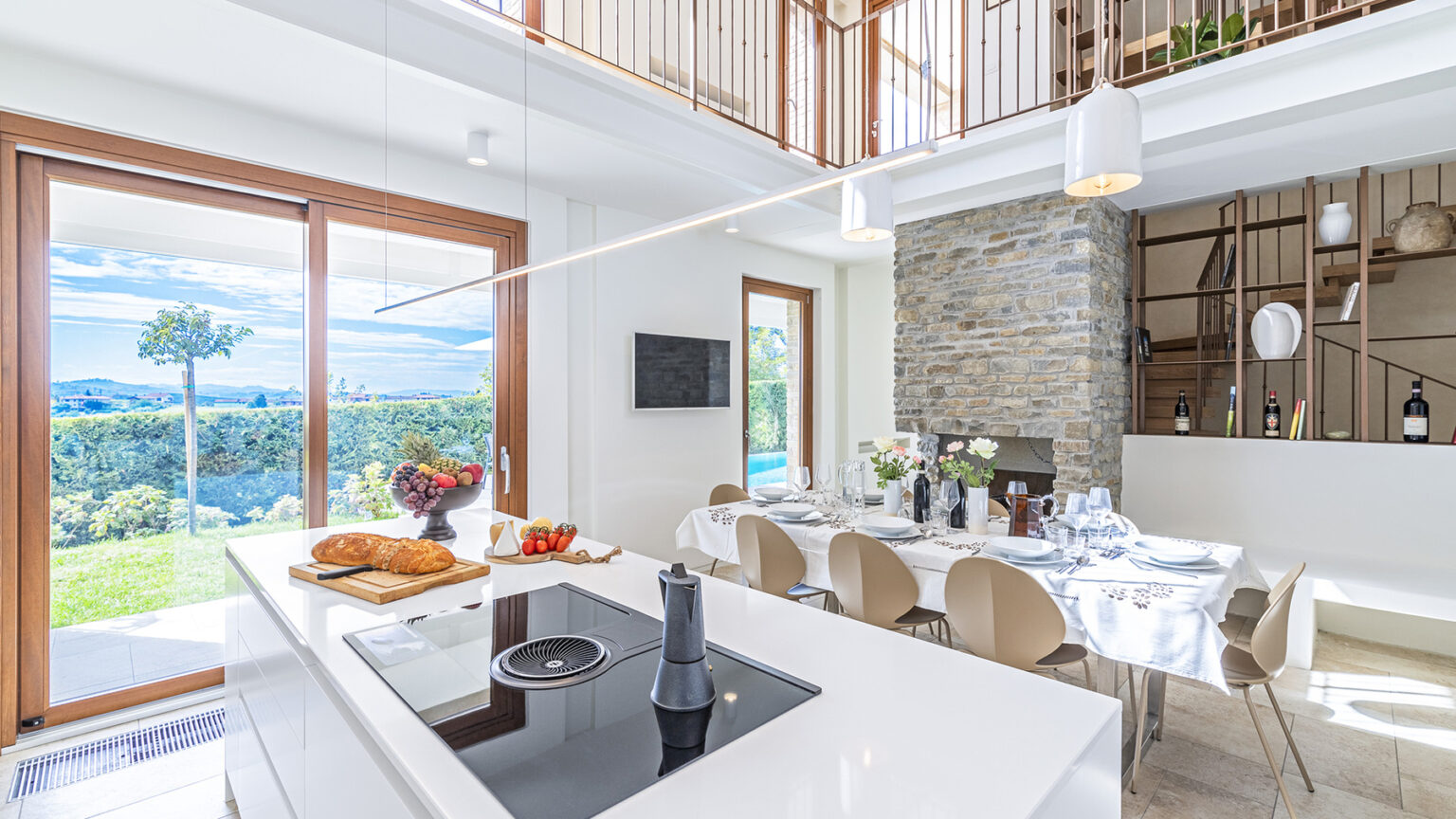 Modern kitchen interior with a sleek white island, an induction cooktop, and a dining area featuring elegant chairs and a stone fireplace. Large windows showcase a scenic outdoor view, enhancing the bright, airy atmosphere of the space. | Only on SopranoVillas