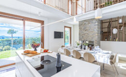 Modern kitchen interior with a sleek white island, an induction cooktop, and a dining area featuring elegant chairs and a stone fireplace. Large windows showcase a scenic outdoor view, enhancing the bright, airy atmosphere of the space.
