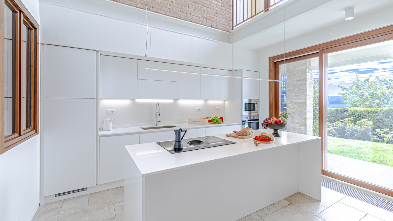 Modern kitchen design featuring a sleek white island, built-in appliances, and large windows providing natural light. The space includes a stylish countertop, fresh fruit, and a view of the outdoors, highlighting a contemporary and functional aesthetic. | Only on SopranoVillas