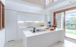 Modern kitchen design featuring a sleek white island, built-in appliances, and large windows providing natural light. The space includes a stylish countertop, fresh fruit, and a view of the outdoors, highlighting a contemporary and functional aesthetic.