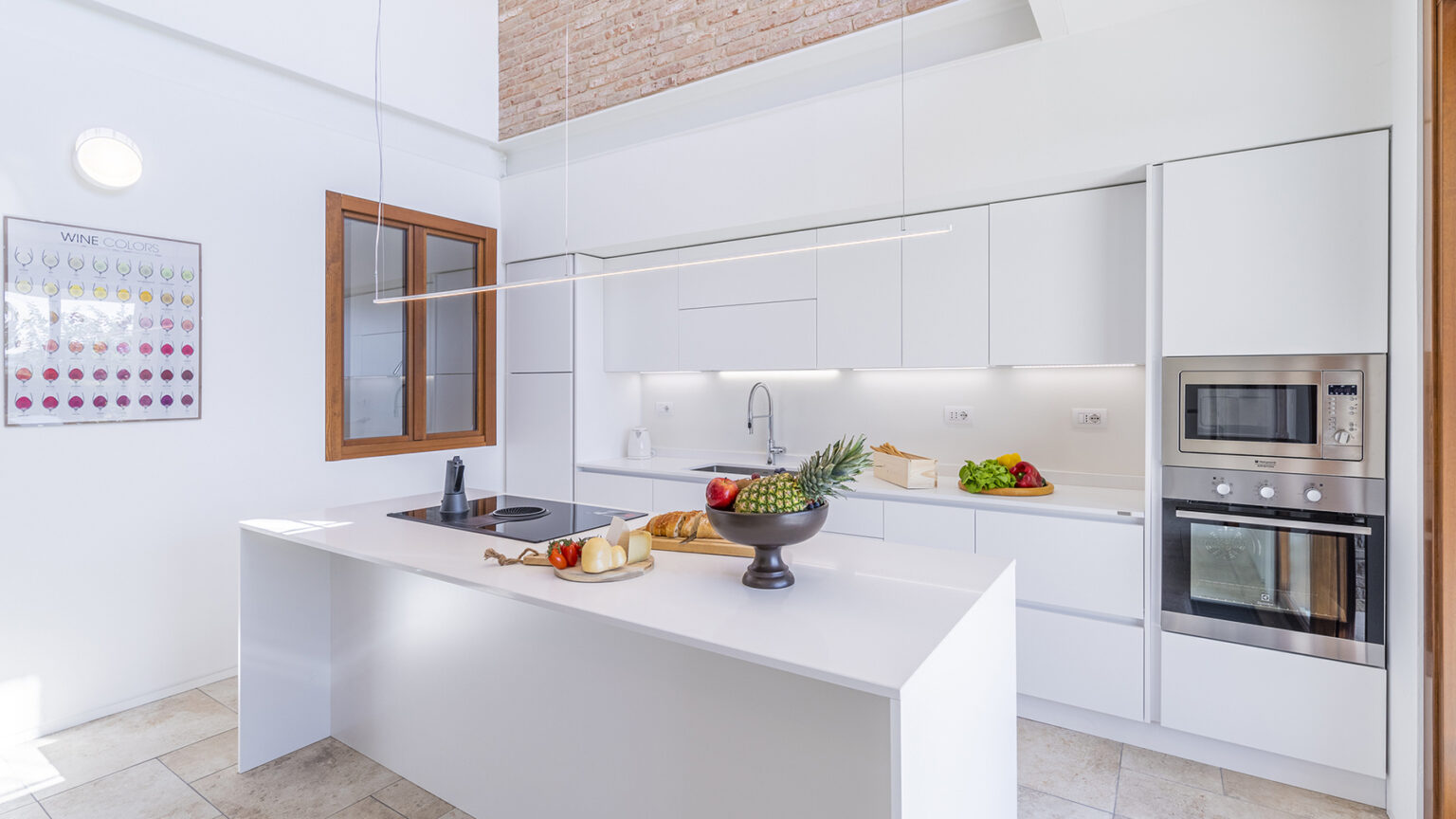 Modern kitchen interior featuring sleek white cabinetry, a central island with a fruit bowl, and built-in ovens. Natural light streams in through large windows, highlighting the spacious design and contemporary decor. | Only on SopranoVillas