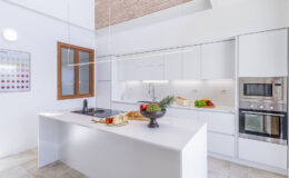 Modern kitchen interior featuring sleek white cabinetry, a central island with a fruit bowl, and built-in ovens. Natural light streams in through large windows, highlighting the spacious design and contemporary decor.