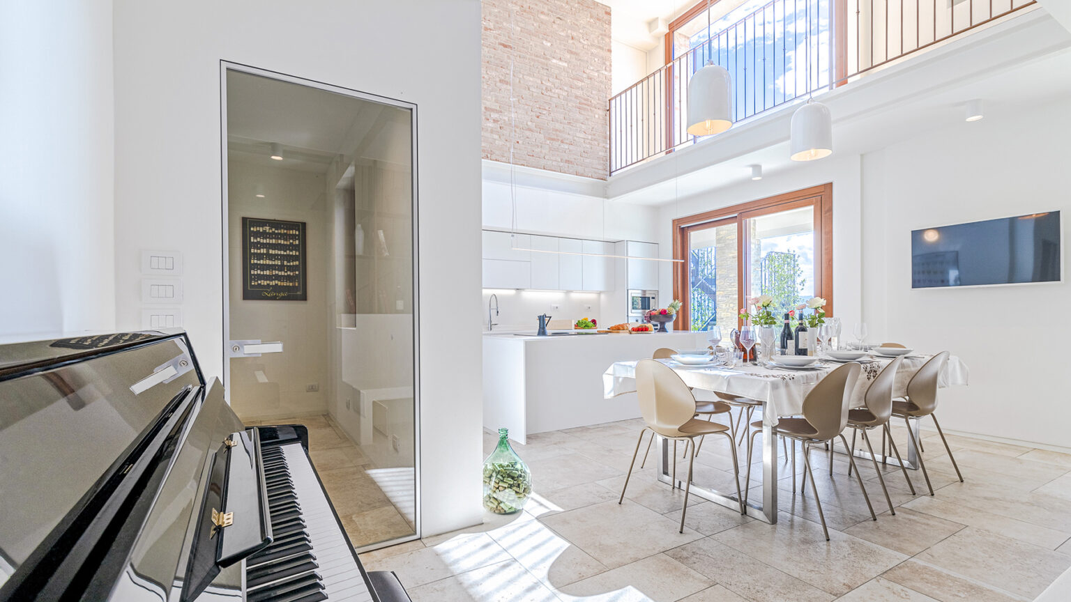 Modern open-concept kitchen and dining area featuring a piano, stylish furniture, and abundant natural light, with a view of a balcony and an adjacent bathroom. | Only on SopranoVillas
