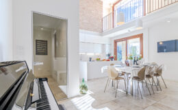 Modern open-concept kitchen and dining area featuring a piano, stylish furniture, and abundant natural light, with a view of a balcony and an adjacent bathroom.
