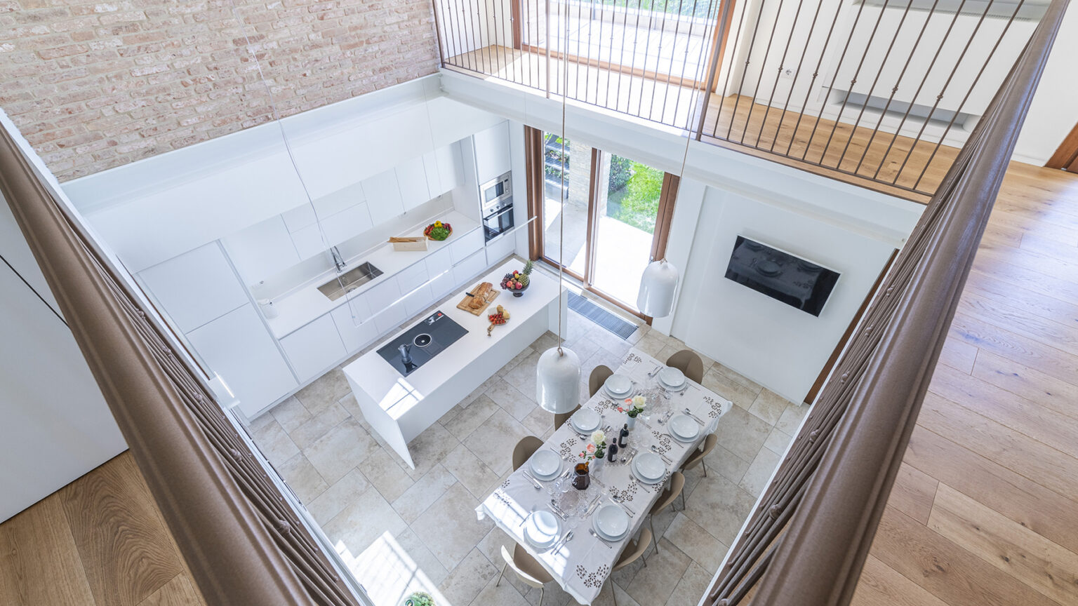 Modern kitchen interior with a spacious layout, featuring white cabinetry, a central island with a cooktop, and a dining area set for a meal. Large windows provide natural light and a view of the garden, complemented by stylish pendant lights and a wooden floor. | Only on SopranoVillas