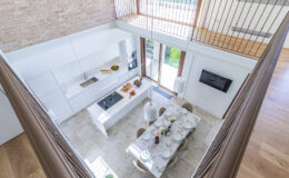 Modern kitchen interior with a spacious layout, featuring white cabinetry, a central island with a cooktop, and a dining area set for a meal. Large windows provide natural light and a view of the garden, complemented by stylish pendant lights and a wooden floor.