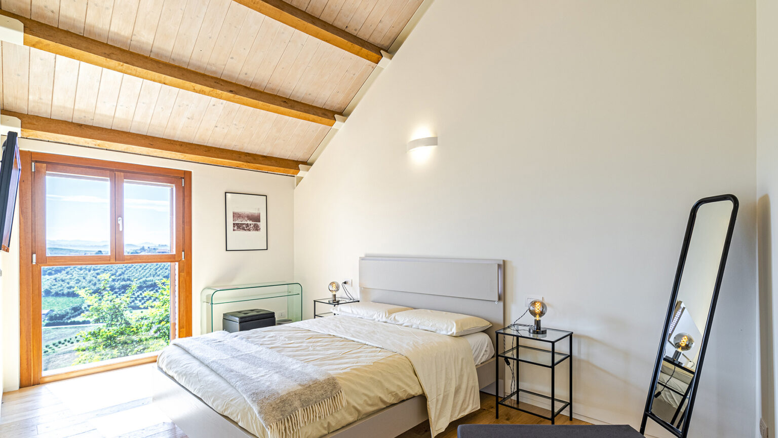 Modern bedroom with a sloped wooden ceiling, featuring a queen-sized bed with neutral bedding, a large window offering scenic views, a stylish floor mirror, and contemporary bedside tables with decorative lamps. | Only on SopranoVillas