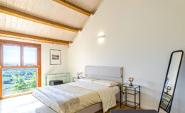 Modern bedroom with a sloped wooden ceiling, featuring a queen-sized bed with neutral bedding, a large window offering scenic views, a stylish floor mirror, and contemporary bedside tables with decorative lamps.