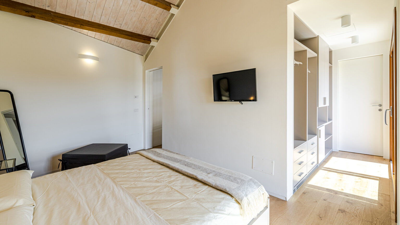 Cozy bedroom featuring a comfortable bed with a light-colored duvet, wooden beams on the ceiling, a wall-mounted TV, and a spacious closet area with drawers. Natural light illuminates the room, creating a warm and inviting atmosphere. | Only on SopranoVillas