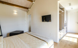 Cozy bedroom featuring a comfortable bed with a light-colored duvet, wooden beams on the ceiling, a wall-mounted TV, and a spacious closet area with drawers. Natural light illuminates the room, creating a warm and inviting atmosphere.