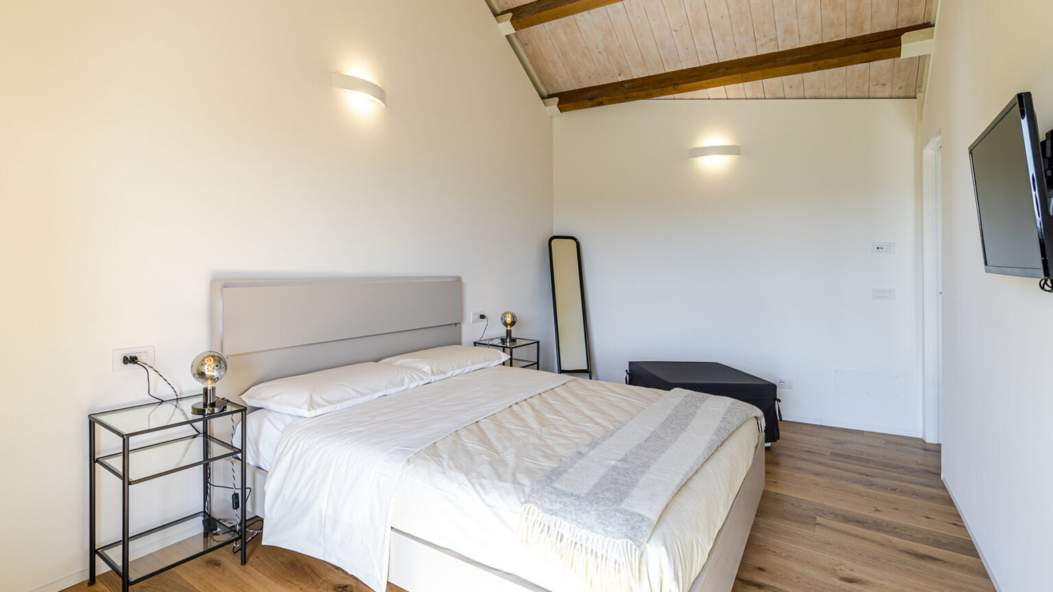 Modern bedroom with a queen-sized bed, wooden ceiling beams, stylish bedside tables, a full-length mirror, and a wall-mounted TV, featuring neutral colors and natural light. | Only on SopranoVillas