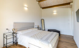 Modern bedroom with a queen-sized bed, wooden ceiling beams, stylish bedside tables, a full-length mirror, and a wall-mounted TV, featuring neutral colors and natural light.