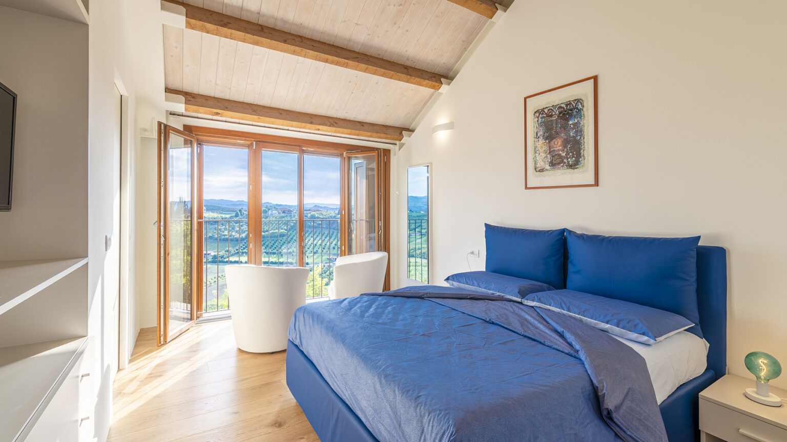 Bright and modern bedroom featuring a blue bedspread, large windows with scenic views, and minimalist decor. The room includes a stylish chair, a mirror, and a bedside lamp, all set against a backdrop of wooden beams and natural light. | Only on SopranoVillas
