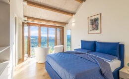 Bright and modern bedroom featuring a blue bedspread, large windows with scenic views, and minimalist decor. The room includes a stylish chair, a mirror, and a bedside lamp, all set against a backdrop of wooden beams and natural light.
