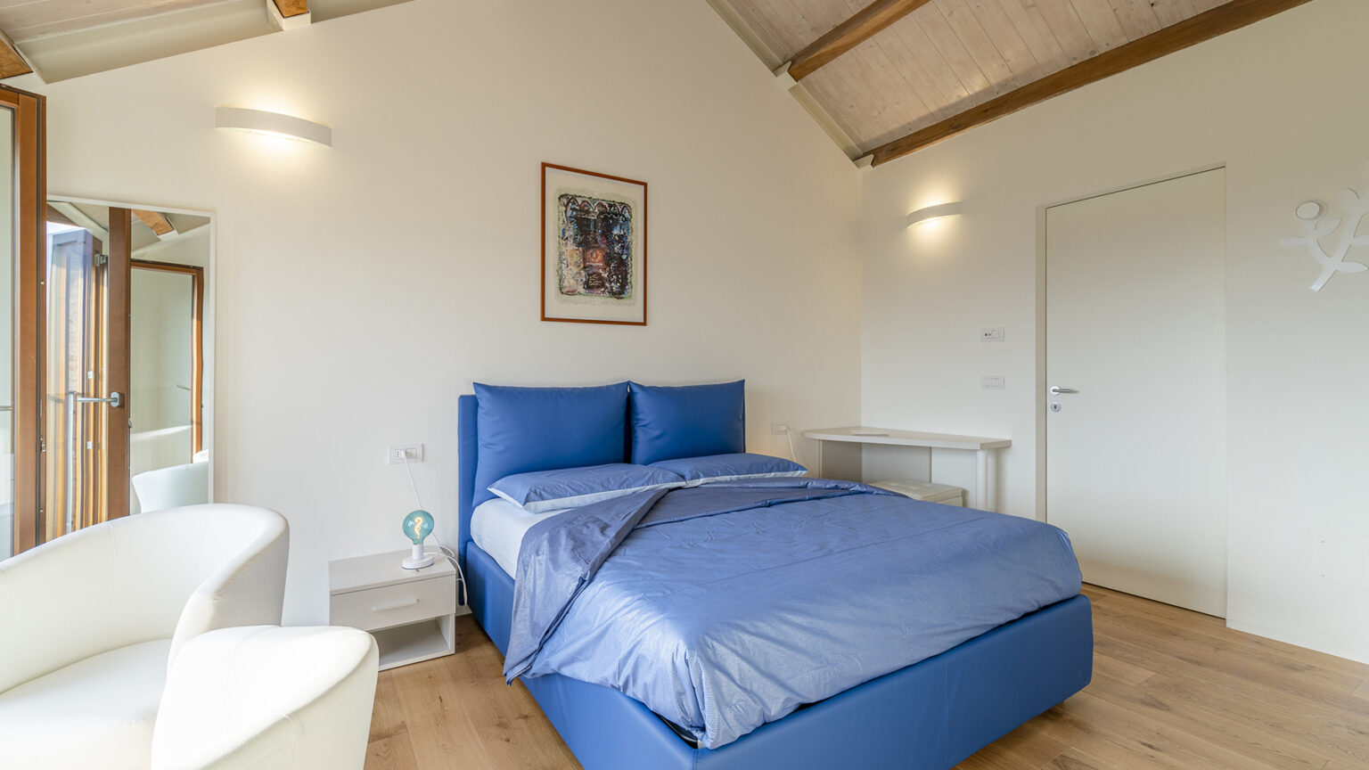 Modern bedroom featuring a blue bed with matching linens, a white armchair, and a minimalist desk. The room has wooden beams, natural light from large windows, and a contemporary artwork on the wall, creating a cozy and stylish atmosphere. | Only on SopranoVillas