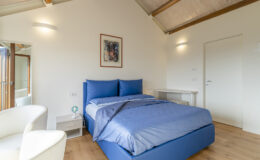 Modern bedroom featuring a blue bed with matching linens, a white armchair, and a minimalist desk. The room has wooden beams, natural light from large windows, and a contemporary artwork on the wall, creating a cozy and stylish atmosphere.