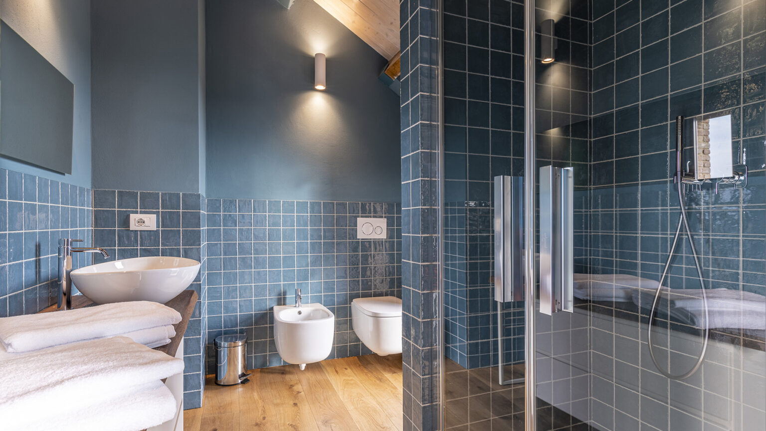 Modern bathroom featuring blue tiled walls, a glass shower enclosure, a bidet, and a stylish sink with neatly arranged towels, showcasing a blend of functionality and contemporary design. | Only on SopranoVillas