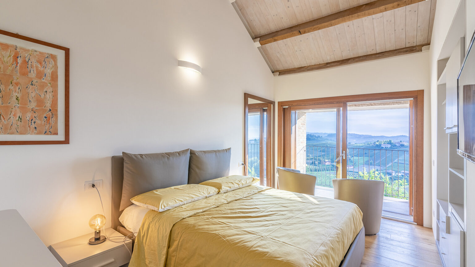 Modern bedroom with a yellow bedspread, large windows offering a scenic view, and a cozy atmosphere. The room features wooden beams, minimalistic decor, and bedside lighting, perfect for relaxation. | Only on SopranoVillas