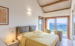 Modern bedroom with a yellow bedspread, large windows offering a scenic view, and a cozy atmosphere. The room features wooden beams, minimalistic decor, and bedside lighting, perfect for relaxation.