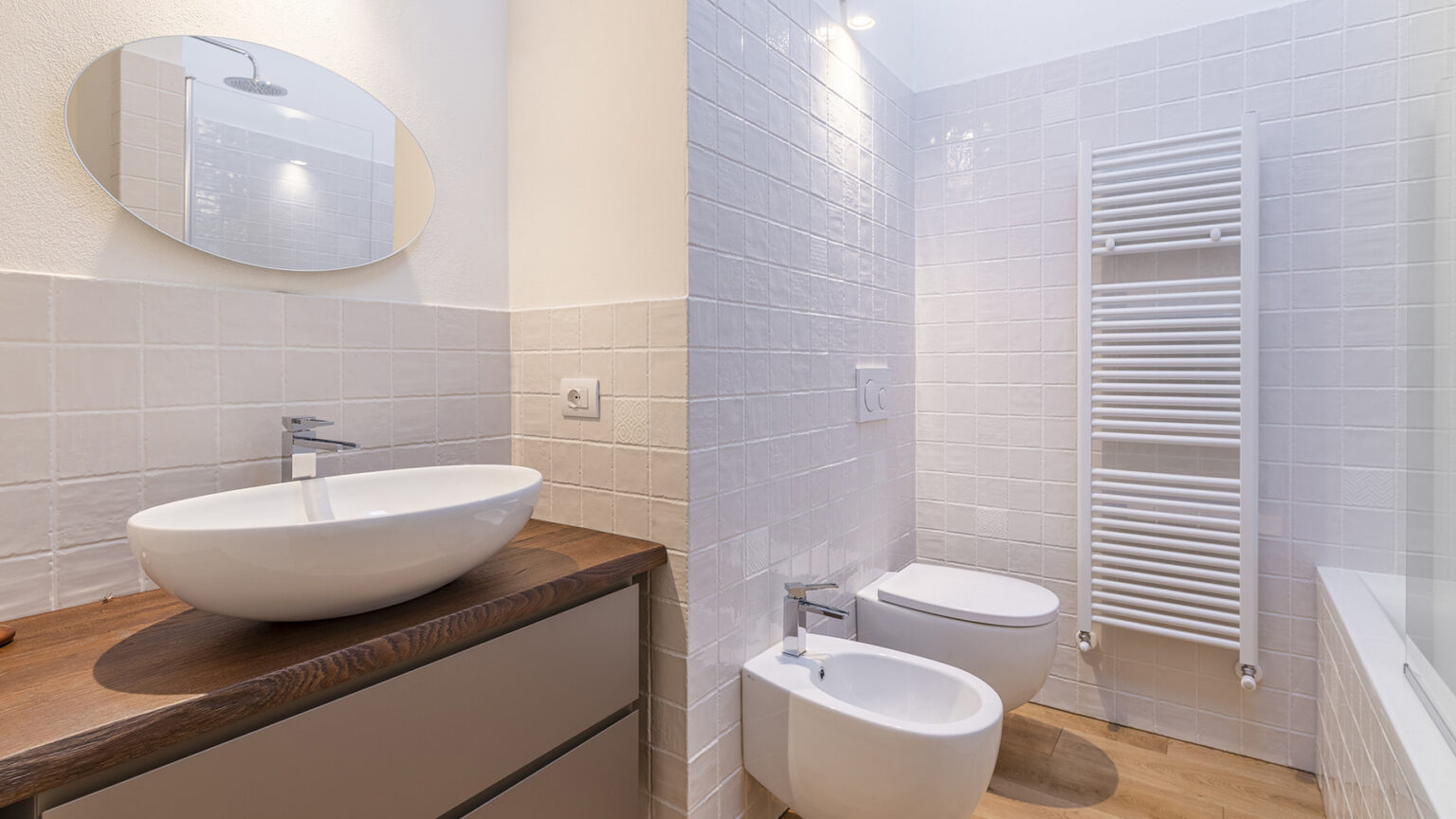 Modern bathroom design featuring a sleek round mirror, a stylish vessel sink on a wooden countertop, a bidet, and a towel radiator against white tiled walls. | Only on SopranoVillas