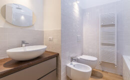 Modern bathroom design featuring a sleek round mirror, a stylish vessel sink on a wooden countertop, a bidet, and a towel radiator against white tiled walls.