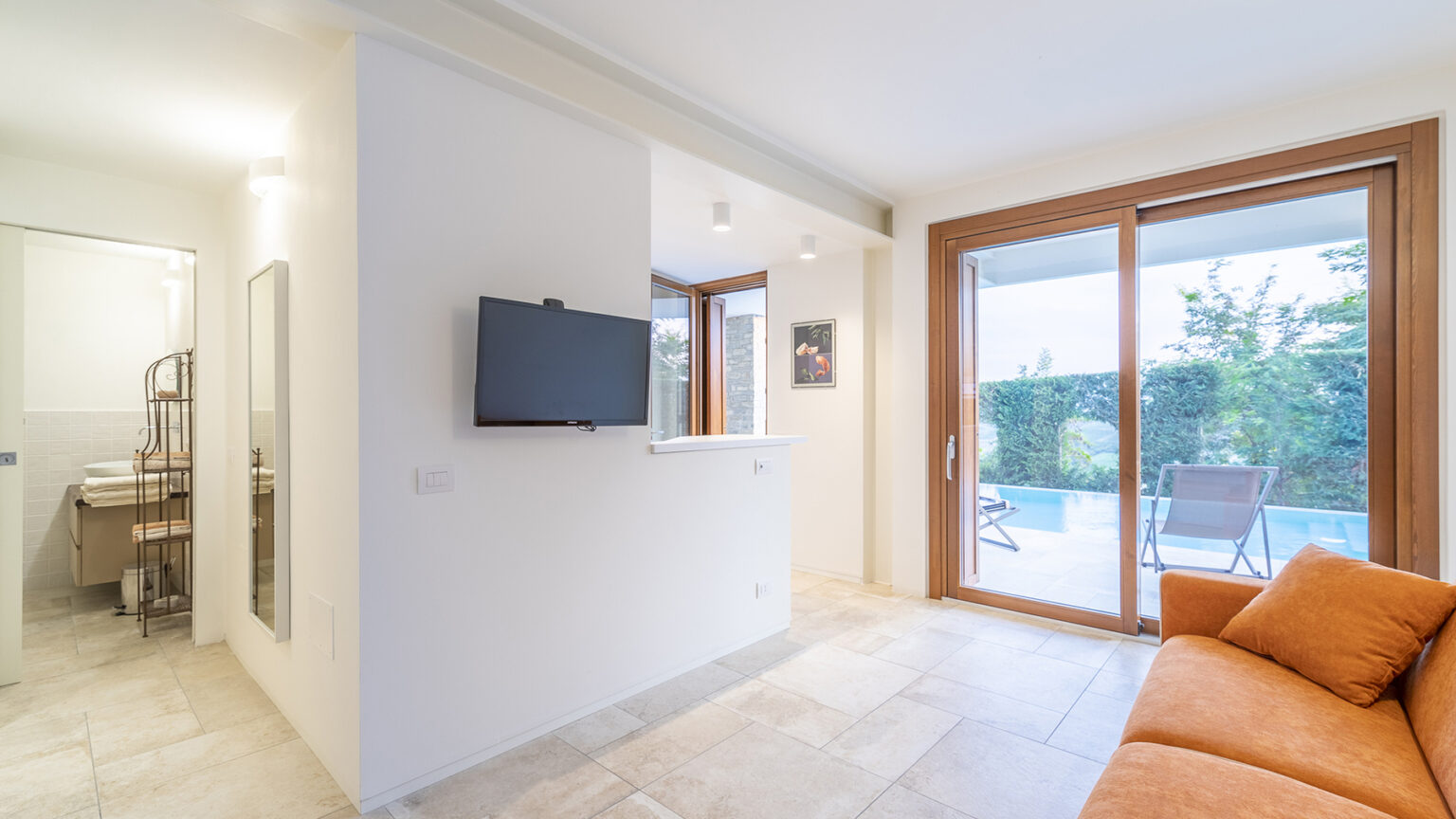 Modern living room with an orange sofa, a wall-mounted TV, and large glass doors leading to an outdoor patio. The space features neutral colors and natural light, creating a cozy atmosphere. | Only on SopranoVillas